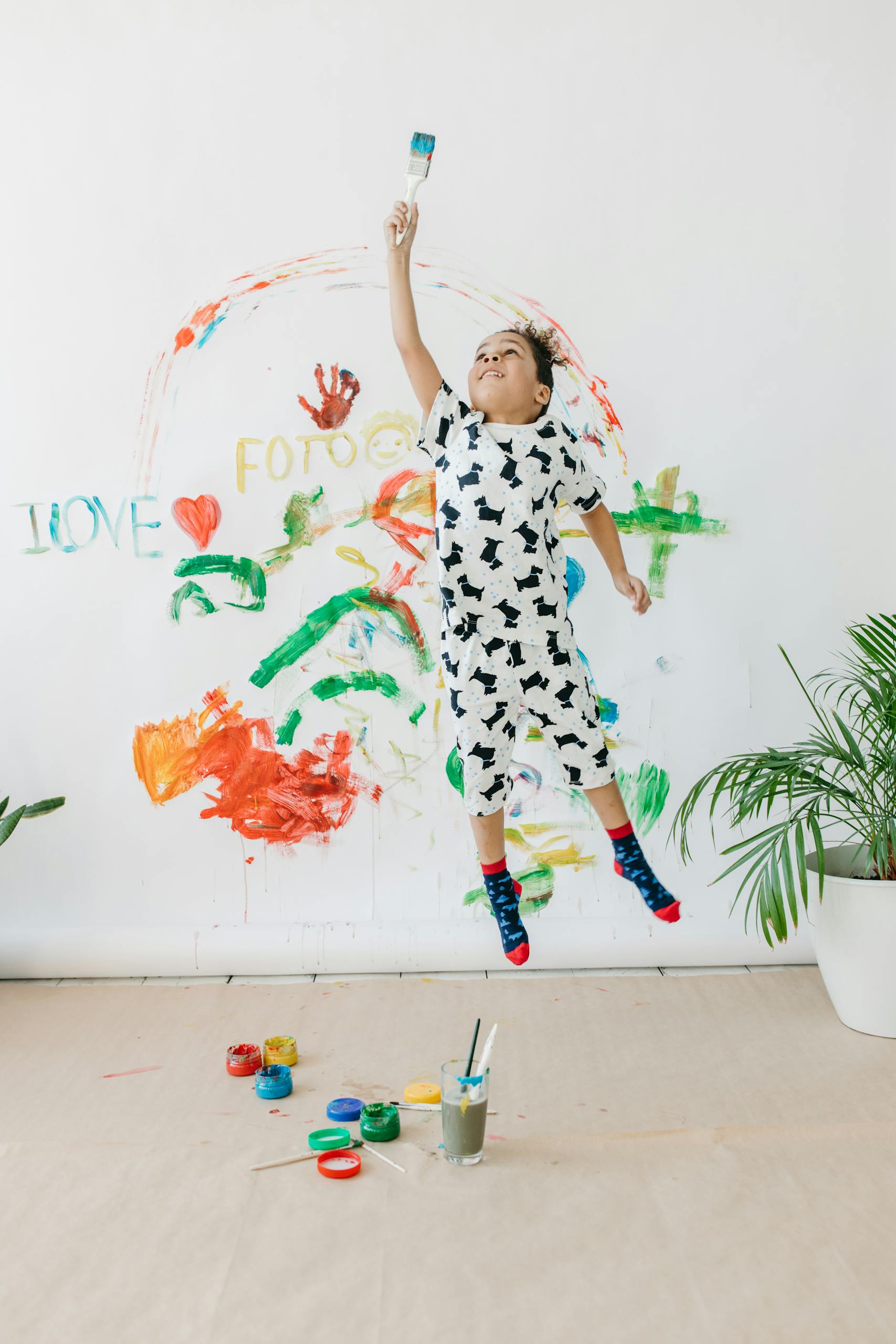 A playful child joyfully jumps holding a paintbrush, creating vibrant wall art with colorful paint.